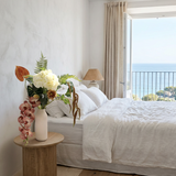 NO. 70 by Nicolíe de Luxe: ivory hydrangea, terracotta anthurium, cascading blush orchids, fern and trailing amaranthus in a white bottle vase, styled on an oak bedside table in a Marbella villa bedroom with sea view