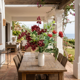 NO. 72 by Nicolíe de Luxe: burgundy berry clusters, red chrysanthemums, a central protea, ferns and trailing amaranthus in a white bottle vase, styled on an oak dining table on a covered Marbella villa terrace