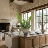 NO. 43 by Nicolíe de Luxe: green bells, olive branches and hanging green amaranthus in a fluted smoky-mauve glass vase, styled on a rustic oak sideboard in a stone-walled villa living room
