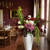 NO. 62 by Nicolíe de Luxe: blush hydrangea, cream protea and ivory roses with burgundy berries, green bells and trailing amaranthus in a white vase, styled on a walnut sideboard in a burgundy villa dining room