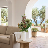 NO. 65 by Nicolíe de Luxe: green anemones, brown-purple orchids, caramel blooms, berries and eucalyptus in a white bottle vase, styled on a travertine side table in a Marbella villa living room with sea-view terrace