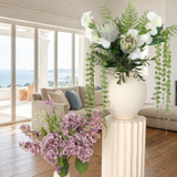 Villa Package NO. 1 by Nicolíe de Luxe: white blooms, proteas, ferns and trailing greenery on a fluted cream pedestal with a lilac hydrangea bouquet beside it, styled in a Marbella villa living room with sea view