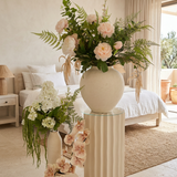 Villa Package NO. 3 by Nicolíe de Luxe: blush peonies and ferns on a fluted cream pedestal with white hydrangea and amaranthus and cascading blush orchids beside it, styled in a Marbella villa bedroom