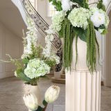 Villa Package NO. 4 by Nicolíe de Luxe: green hydrangeas, white peonies and trailing amaranthus on a fluted cream pedestal with white delphinium and proteas beside it, styled in a Marbella villa entrance foyer