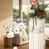 Villa Package NO. 5 by Nicolíe de Luxe: blush orchids, ivory and bronze hydrangeas with trailing greenery on a fluted cream pedestal beside white magnolia branches, styled in a modern Marbella villa lounge with pool view